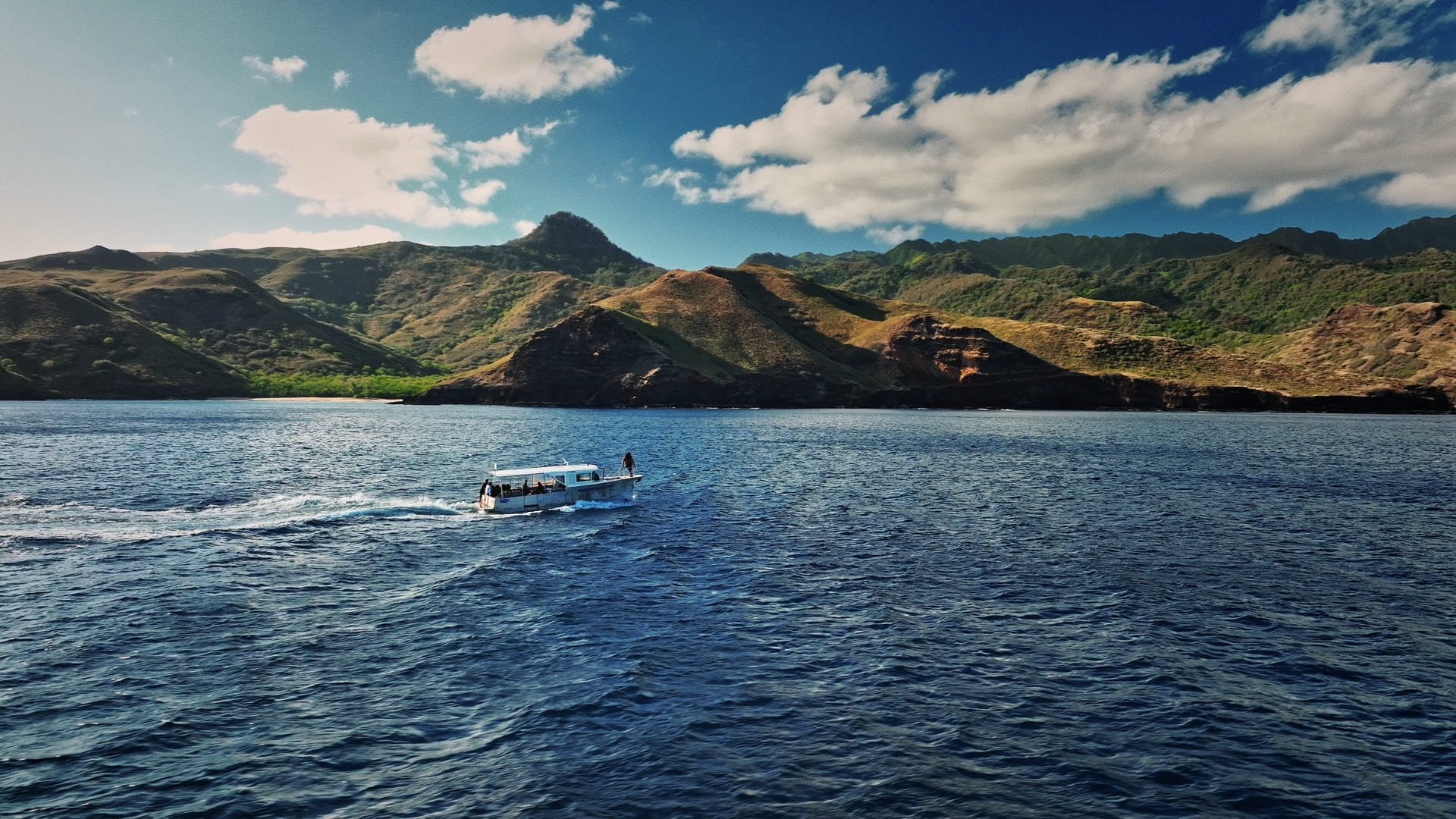 Small boat sailing on a blue sea in front of green mountains under a partly cloudy sky.